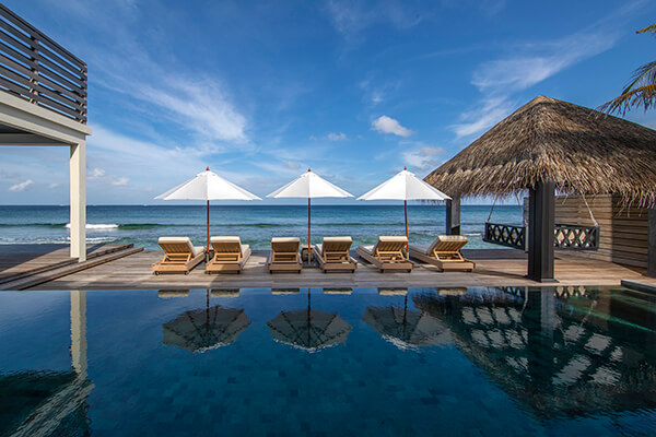 An exotic tropical landscape with a clear blue sky. There is a wooden deck with a small seating area, consisting of lounge chaises. The chairs face the ocean, with a white umbrella providing shade.