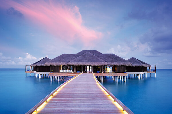 A lit up wooden deck over the water leading to hut style rooms with beautiful tropical island landscape. The water is crystal-clear. The overall image conveys a sense of peacefulness, tranquility, and the beauty of nature. 